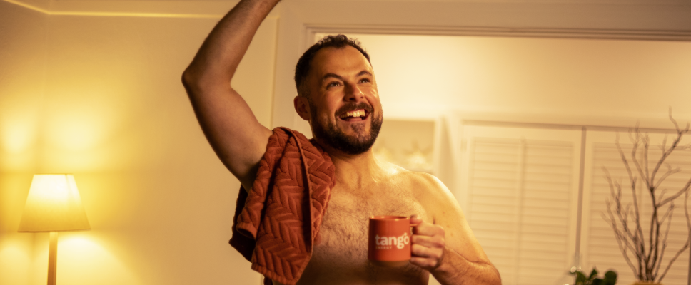 A man smiling and looking up. He is shirtless and is standing in front of a basket of laundry. One hand is in the air and one hand is holding a mug.